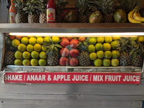 A fruit display featuring neatly arranged rows of pineapples, citrus fruit, pomegranates, and a few bananas on the side. Two juice bottles are also visible on the shelf. A sign below advertises various fruit juices like Shake, Anaar (pomegranate), Apple Juice, and Mix Fruit Juice.