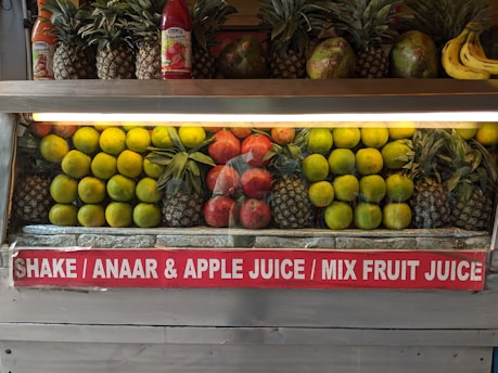 A fruit display featuring neatly arranged rows of pineapples, citrus fruit, pomegranates, and a few bananas on the side. Two juice bottles are also visible on the shelf. A sign below advertises various fruit juices like Shake, Anaar (pomegranate), Apple Juice, and Mix Fruit Juice.