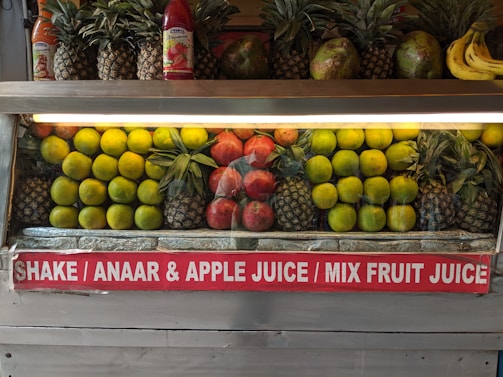A fruit display featuring neatly arranged rows of pineapples, citrus fruit, pomegranates, and a few bananas on the side. Two juice bottles are also visible on the shelf. A sign below advertises various fruit juices like Shake, Anaar (pomegranate), Apple Juice, and Mix Fruit Juice.
