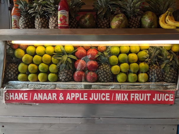 A fruit display featuring neatly arranged rows of pineapples, citrus fruit, pomegranates, and a few bananas on the side. Two juice bottles are also visible on the shelf. A sign below advertises various fruit juices like Shake, Anaar (pomegranate), Apple Juice, and Mix Fruit Juice.