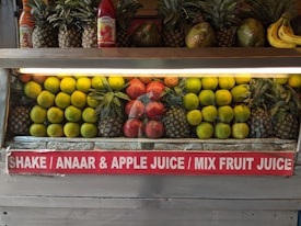 A fruit display featuring neatly arranged rows of pineapples, citrus fruit, pomegranates, and a few bananas on the side. Two juice bottles are also visible on the shelf. A sign below advertises various fruit juices like Shake, Anaar (pomegranate), Apple Juice, and Mix Fruit Juice.
