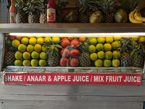 A fruit display featuring neatly arranged rows of pineapples, citrus fruit, pomegranates, and a few bananas on the side. Two juice bottles are also visible on the shelf. A sign below advertises various fruit juices like Shake, Anaar (pomegranate), Apple Juice, and Mix Fruit Juice.