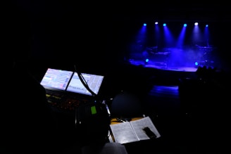 A dimly lit stage is illuminated by blue spotlights, creating an atmospheric setting with visible smoke. In the foreground, a mixing console with lit screens and sound equipment is visible, along with scattered papers and what appears to be a smartphone.