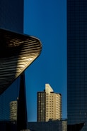 A modern urban scene featuring a mix of architectural styles and elements. The central focus is a tall residential building with a facade of windows, framed by two towering structures with reflective glass surfaces. The edge of a metal construction juts out prominently to the left, creating dynamic curves against the clear blue sky.