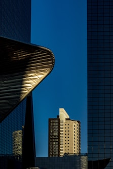 A modern urban scene featuring a mix of architectural styles and elements. The central focus is a tall residential building with a facade of windows, framed by two towering structures with reflective glass surfaces. The edge of a metal construction juts out prominently to the left, creating dynamic curves against the clear blue sky.