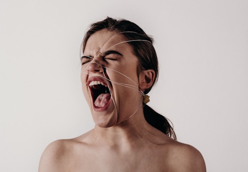 A person with their mouth wide open as if screaming or expressing intense emotion. Their hair is tied back, and there are thin lines crisscrossing their face against a plain background.
