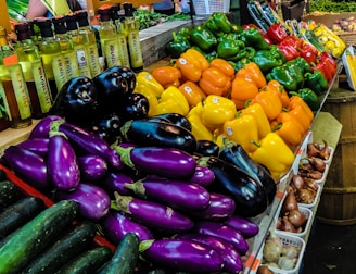 A colorful display of fresh vegetables at a market, including stacks of bell peppers in green, yellow, orange, and red, alongside shiny eggplants and cucumbers. Bottles of oil are seen lined up to the side, with some onions in small baskets. The setting suggests a vibrant and lively produce section.