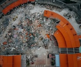 An aerial view of a demolition site with scattered debris and construction equipment. The remains of a building with red-orange roofs surround the area, indicating significant destruction.
