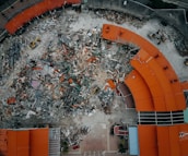 An aerial view of a demolition site with scattered debris and construction equipment. The remains of a building with red-orange roofs surround the area, indicating significant destruction.