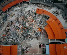 An aerial view of a demolition site with scattered debris and construction equipment. The remains of a building with red-orange roofs surround the area, indicating significant destruction.