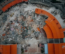 An aerial view of a demolition site with scattered debris and construction equipment. The remains of a building with red-orange roofs surround the area, indicating significant destruction.