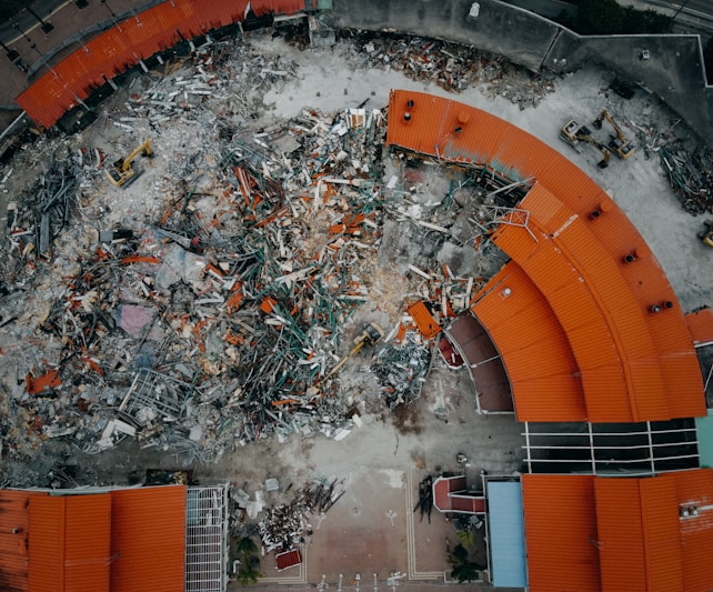 An aerial view of a demolition site with scattered debris and construction equipment. The remains of a building with red-orange roofs surround the area, indicating significant destruction.