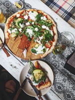 A wooden table is set with a round pizza topped with fresh vegetables like zucchini, spinach, and arugula, as well as dollops of white cheese. A slice has been placed on a separate plate with a knife and fork beside it. There are items such as a drink, likely beer, and some olive oil nearby on a lace tablecloth.