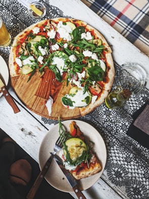 A wooden table is set with a round pizza topped with fresh vegetables like zucchini, spinach, and arugula, as well as dollops of white cheese. A slice has been placed on a separate plate with a knife and fork beside it. There are items such as a drink, likely beer, and some olive oil nearby on a lace tablecloth.