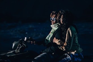 A couple is riding an ATV in a dimly lit environment. The man in front is wearing sunglasses and a scarf, while the woman behind him is holding onto him tightly, dressed in casual clothing. They appear to be in motion, suggesting an adventurous setting, possibly at night.