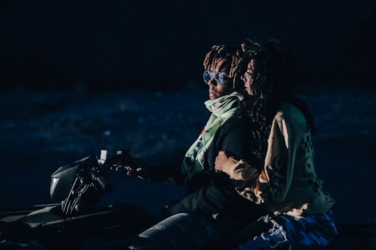 A couple is riding an ATV in a dimly lit environment. The man in front is wearing sunglasses and a scarf, while the woman behind him is holding onto him tightly, dressed in casual clothing. They appear to be in motion, suggesting an adventurous setting, possibly at night.