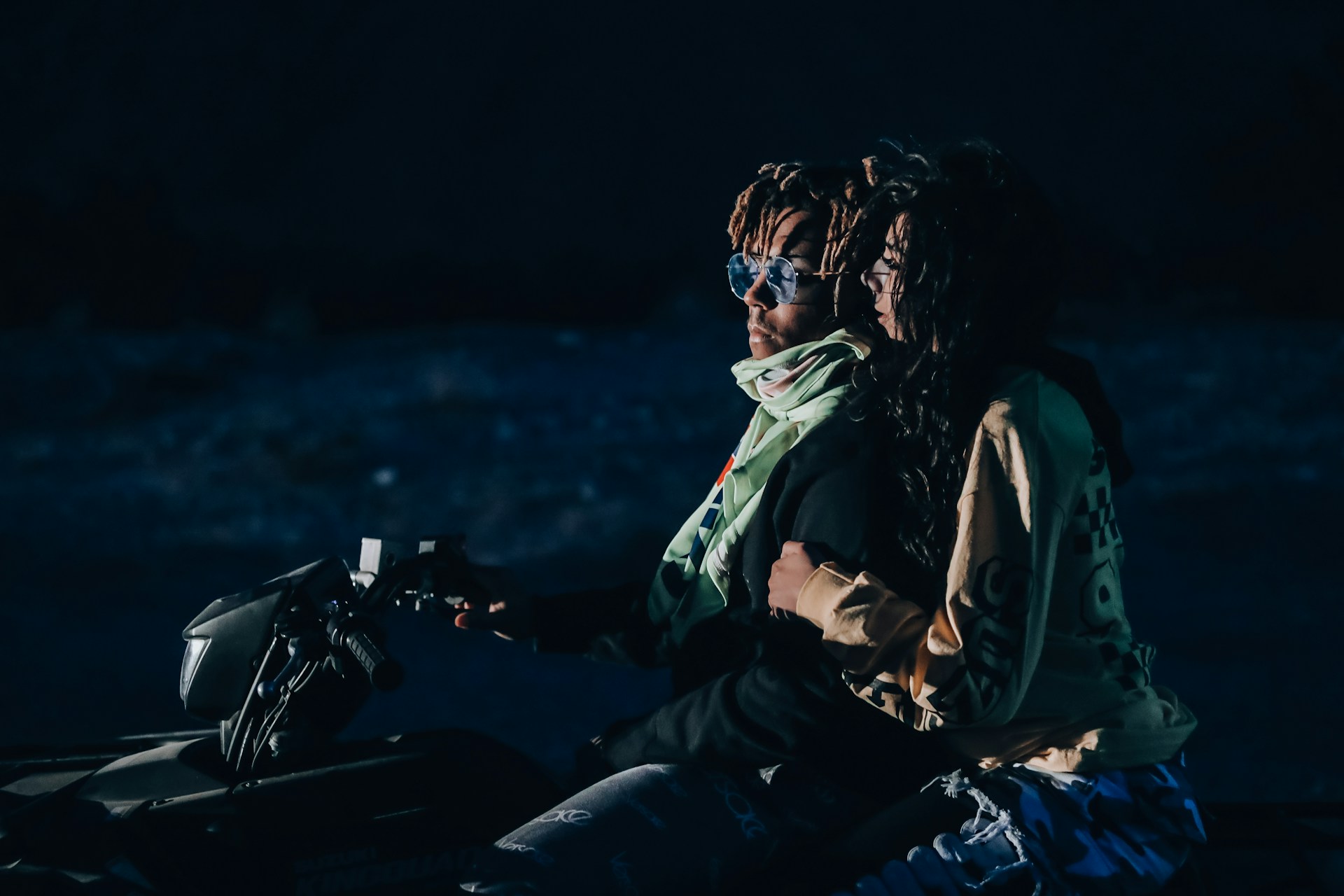 A couple is riding an ATV in a dimly lit environment. The man in front is wearing sunglasses and a scarf, while the woman behind him is holding onto him tightly, dressed in casual clothing. They appear to be in motion, suggesting an adventurous setting, possibly at night.