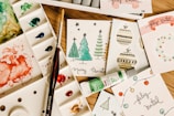 A collection of handmade holiday greeting cards featuring Christmas-themed illustrations, including trees and ornaments, arranged on a wooden surface. Accompanied by a watercolor palette with various colors, a paintbrush, and a tube of green watercolor paint.