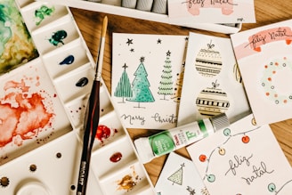 A collection of handmade holiday greeting cards featuring Christmas-themed illustrations, including trees and ornaments, arranged on a wooden surface. Accompanied by a watercolor palette with various colors, a paintbrush, and a tube of green watercolor paint.