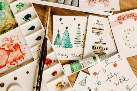 A collection of handmade holiday greeting cards featuring Christmas-themed illustrations, including trees and ornaments, arranged on a wooden surface. Accompanied by a watercolor palette with various colors, a paintbrush, and a tube of green watercolor paint.