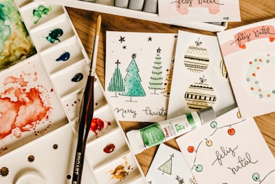 A collection of handmade holiday greeting cards featuring Christmas-themed illustrations, including trees and ornaments, arranged on a wooden surface. Accompanied by a watercolor palette with various colors, a paintbrush, and a tube of green watercolor paint.