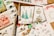 A collection of handmade holiday greeting cards featuring Christmas-themed illustrations, including trees and ornaments, arranged on a wooden surface. Accompanied by a watercolor palette with various colors, a paintbrush, and a tube of green watercolor paint.