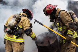 Two firefighters wearing protective gear and helmets are actively engaged in handling a situation involving a barrel amid billowing smoke. One firefighter holds a tool, possibly a hook, as they work together.