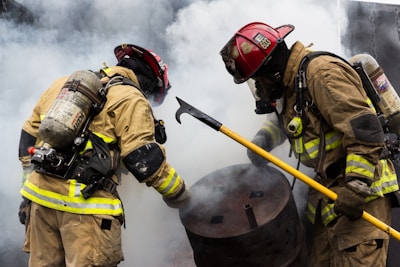 Two firefighters wearing protective gear and helmets are actively engaged in handling a situation involving a barrel amid billowing smoke. One firefighter holds a tool, possibly a hook, as they work together.