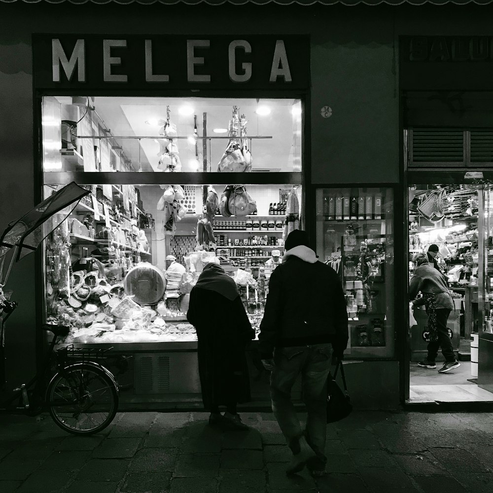 grayscale photo of two person standing on the Melega store