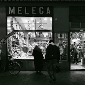 A dimly lit storefront displaying a variety of cured meats and cheeses, with shelves filled with bottles and jars. Two figures are standing outside, appearing to look through the window, while another person is seen working inside.