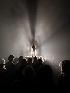 A person stands in a spotlight on a stage, with a large crowd in the foreground shrouded in shadow. Rays of light dramatically emanate from behind the figure, creating a striking silhouette against the illuminated background.