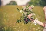 A hand holds a bouquet of flowers, featuring white roses, baby's breath, and lush green leaves. The person is wearing a white lace garment, possibly a wedding dress. The background is a blurred field, suggesting an outdoor setting with green and earthy tones.