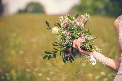 A hand holds a bouquet of flowers, featuring white roses, baby's breath, and lush green leaves. The person is wearing a white lace garment, possibly a wedding dress. The background is a blurred field, suggesting an outdoor setting with green and earthy tones.