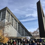 A large group of people is gathered outside a modern church with a distinctive geometric design. The building features intricate patterns on its facade, and a tall, narrow bell tower stands beside it. The scene includes people holding signs, suggesting a demonstration or gathering. Trees with autumn foliage add color to the setting under a blue sky with scattered clouds.