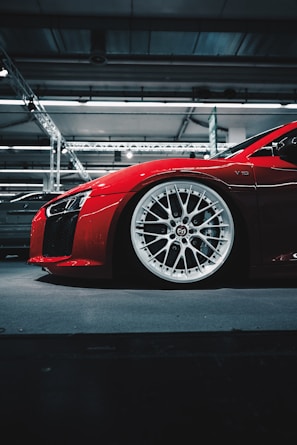 A red sports car showcased indoors, highlighting its sleek design and white, intricate alloy wheels. The reflective surface of the car glistens under the overhead lighting, and the industrial setting with metal beams and lights provides a modern background.