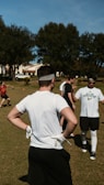 Several people engage in an outdoor sporting activity on a grass field. The central figure is a man in a white shirt, black shorts, and a headband, standing with hands on hips and wearing gloves. Other participants are visible in the background, interacting casually. Green trees and houses line the area under a clear blue sky.