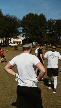 Several people engage in an outdoor sporting activity on a grass field. The central figure is a man in a white shirt, black shorts, and a headband, standing with hands on hips and wearing gloves. Other participants are visible in the background, interacting casually. Green trees and houses line the area under a clear blue sky.