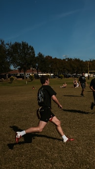 A person is running on a grassy field with several other people in the background. The setting appears to be a park or open recreational area with trees and buildings visible in the distance. The individuals are wearing athletic clothing, suggesting they might be engaging in a sport.