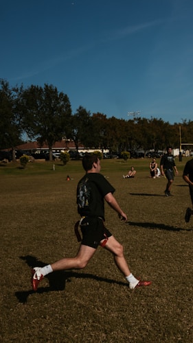 A person is running on a grassy field with several other people in the background. The setting appears to be a park or open recreational area with trees and buildings visible in the distance. The individuals are wearing athletic clothing, suggesting they might be engaging in a sport.