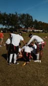 A group of individuals in athletic attire are huddled together on a grass field, possibly strategizing for a flag football game. They wear red and black flag belts and white shirts, with their attention focused inward. The surrounding area shows a sunny day with trees in the background and a clear blue sky.