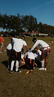A group of individuals in athletic attire are huddled together on a grass field, possibly strategizing for a flag football game. They wear red and black flag belts and white shirts, with their attention focused inward. The surrounding area shows a sunny day with trees in the background and a clear blue sky.