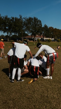 A group of individuals in athletic attire are huddled together on a grass field, possibly strategizing for a flag football game. They wear red and black flag belts and white shirts, with their attention focused inward. The surrounding area shows a sunny day with trees in the background and a clear blue sky.
