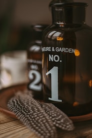 Two vintage brown glass bottles labeled 'NATURE & GARDEN NO. 1' and 'NO. 2' are placed on a terracotta plate with spotted feathers. The composition has a rustic and natural aesthetic.