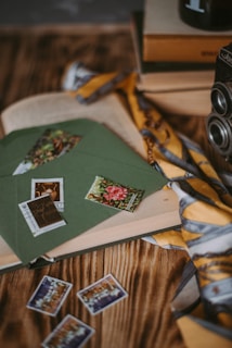 An assortment of vintage items including a stack of books, an old camera, and green envelopes with colorful stamps. A yellow and gray patterned scarf is draped over the scene, and more stamps are spread out on a wooden surface.