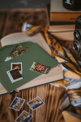 An assortment of vintage items including a stack of books, an old camera, and green envelopes with colorful stamps. A yellow and gray patterned scarf is draped over the scene, and more stamps are spread out on a wooden surface.