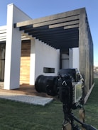 A modern architectural structure with a white facade and wooden accents, featuring black metal beams extending from the roof. A professional DSLR camera mounted on a tripod is focused on the building, highlighting the interaction between architecture and photography.
