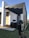 A modern architectural structure with a white facade and wooden accents, featuring black metal beams extending from the roof. A professional DSLR camera mounted on a tripod is focused on the building, highlighting the interaction between architecture and photography.