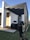 A modern architectural structure with a white facade and wooden accents, featuring black metal beams extending from the roof. A professional DSLR camera mounted on a tripod is focused on the building, highlighting the interaction between architecture and photography.
