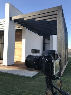 A modern architectural structure with a white facade and wooden accents, featuring black metal beams extending from the roof. A professional DSLR camera mounted on a tripod is focused on the building, highlighting the interaction between architecture and photography.