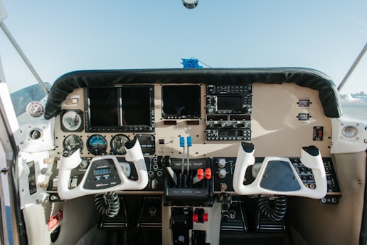 The image depicts the cockpit of a small aircraft, featuring an array of controls and instruments. There are two yokes for steering, various gauges, screens, and switches. The cockpit has a beige and black color scheme, and the exterior view shows a hint of blue sky.